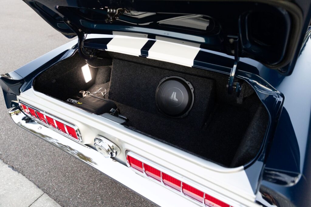 Open trunk of a 1968 Revology Shelby GT500KR showing a custom carpeted interior, LED lighting, and an integrated JL Audio subwoofer enclosure.