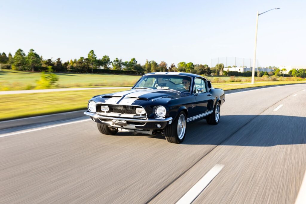 Action shot of a dark green 1968 Revology Shelby GT500KR Fastback driving on a highway, showing the front three-quarter view with white Le Mans stripes.