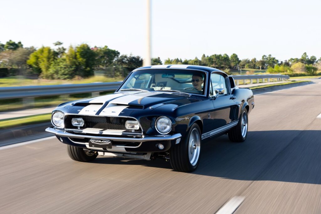 Front three-quarter action shot of a dark green 1968 Revology Shelby GT500KR Fastback with white racing stripes driving on an open highway.