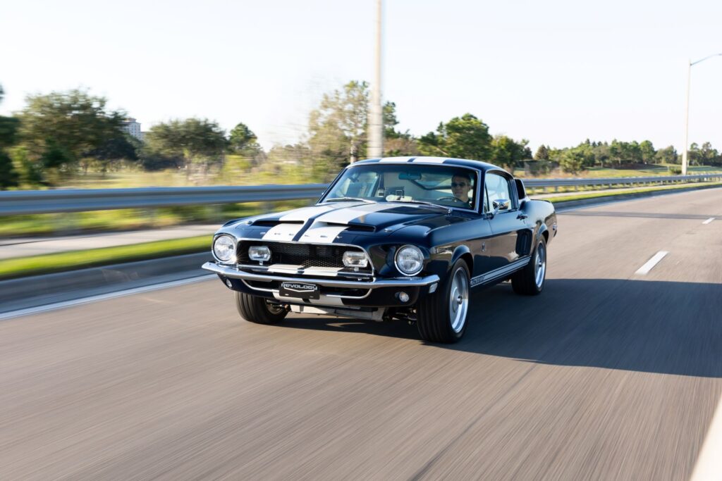 Lower-angle action shot of a dark green 1968 Revology Shelby GT500KR Fastback with white Le Mans stripes driving at speed on a highway.