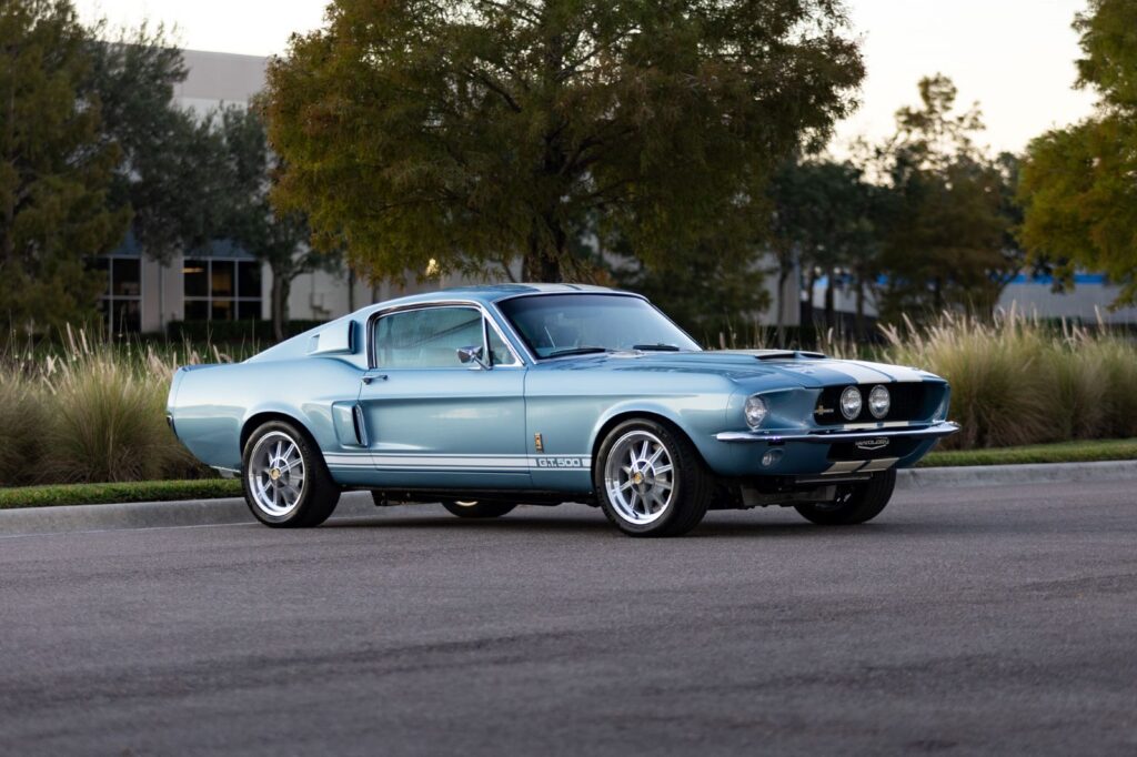 A light blue 1967 Revology Shelby GT500 Fastback with white racing stripes, parked on an asphalt road with trees in the background.