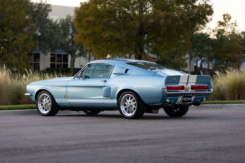 A light blue 1967 Revology Shelby GT500 Fastback restomod with white racing stripes parked on an asphalt road.