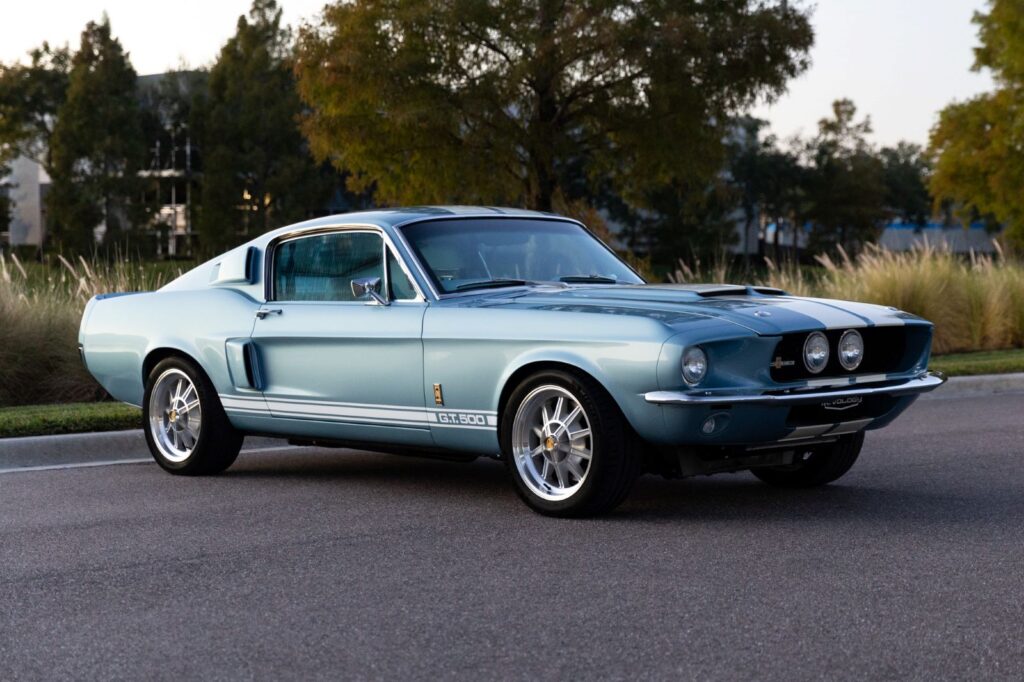 A 1967 Shelby GT500 Fastback by Revology Cars in metallic light blue with white racing stripes, parked on an asphalt road at dusk.