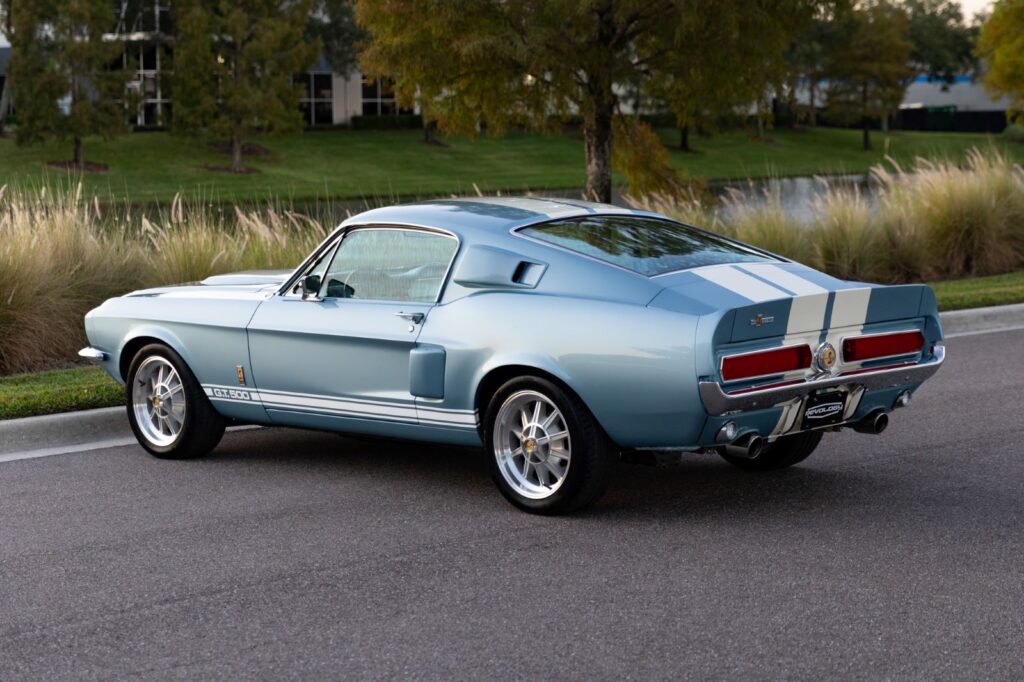 Rear three-quarter view of a metallic blue 1967 Revology Shelby GT500 Fastback showing the sloping roofline, white racing stripes, and signature rear spoiler.
