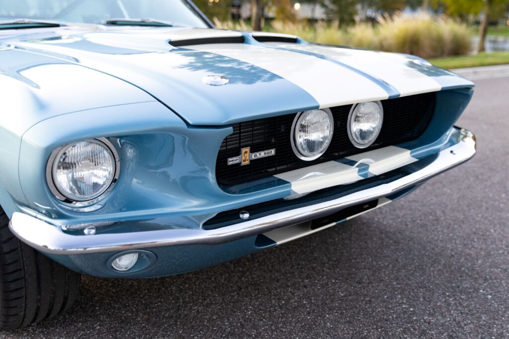 Close-up of the front grille and hood of a metallic blue 1967 Revology Shelby GT500, featuring center-mounted driving lights, white racing stripes, and chrome bumper.