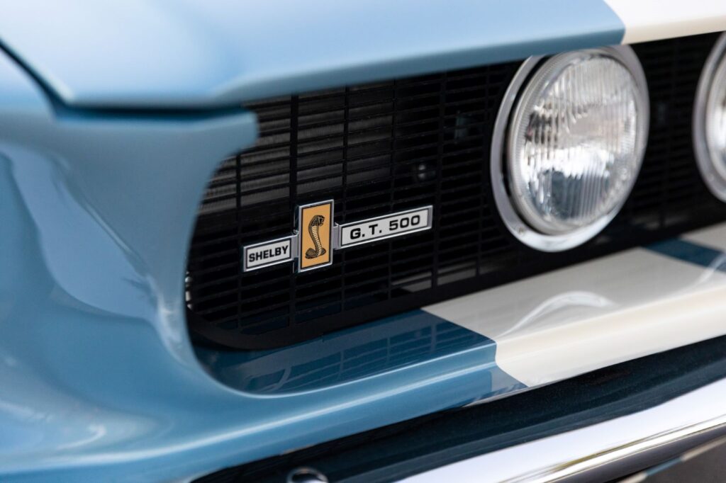 Close-up of the Shelby GT500 Cobra emblem on the black front grille of a 1967 Revology Mustang, featuring white racing stripes and a driving light.