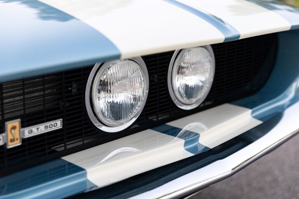 Close-up of the dual center-mounted driving lights on a 1967 Revology Shelby GT500 Fastback with white racing stripes on a metallic blue hood.