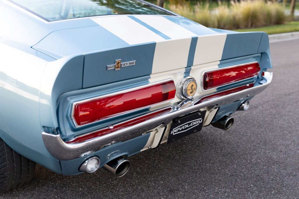 Rear view detail of a metallic blue 1967 Revology Shelby GT500 Fastback showing the horizontal taillights, chrome bumper, and dual exhaust tips.