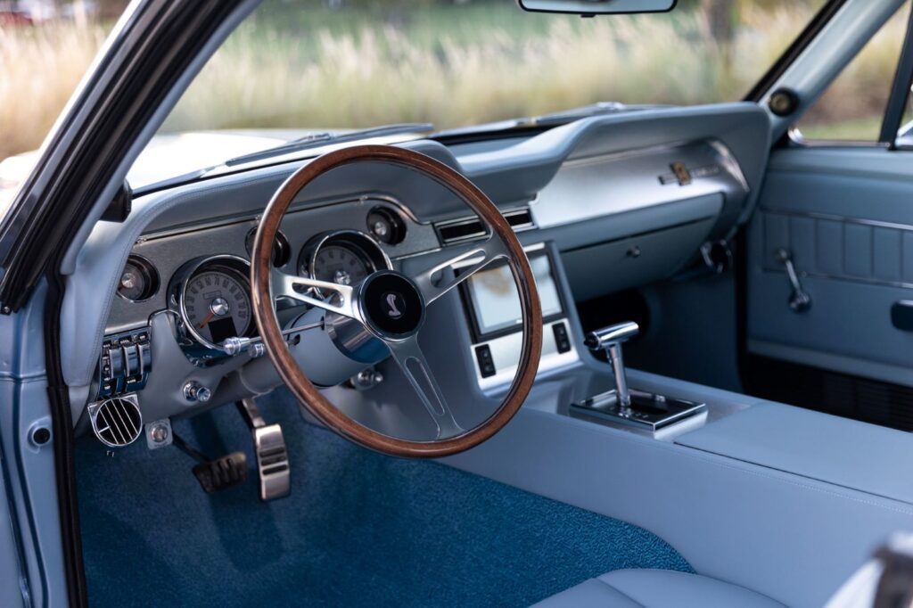Close-up of the light blue leather dashboard in a 1967 Revology Shelby GT500, featuring a wood-rimmed steering wheel, modern electronic gauges, and a touchscreen infotainment system.