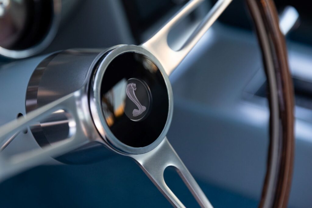 Close-up of a Shelby Cobra emblem on the center horn button of a wood-rimmed three-spoke steering wheel in a 1967 Revology GT500.