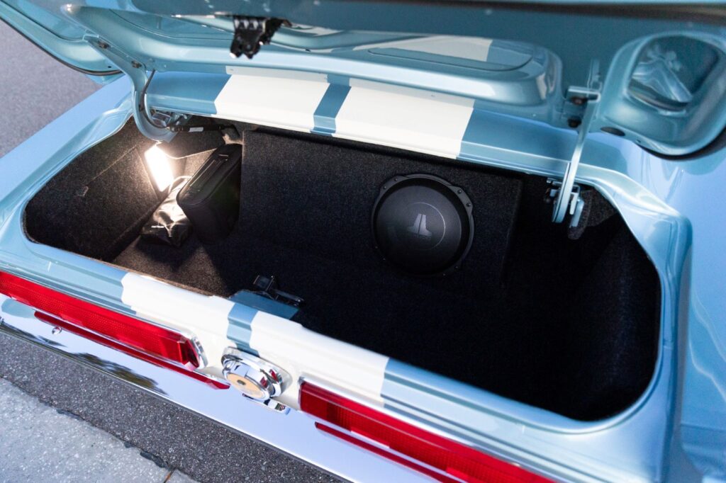 Open trunk of a 1967 Revology Shelby GT500 featuring a custom-fitted JL Audio subwoofer and amplifier integrated into a carpeted enclosure.