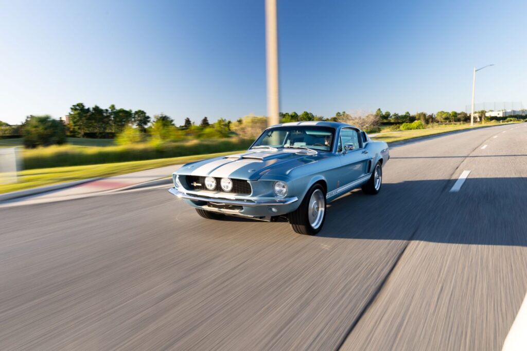 A metallic blue 1967 Revology Shelby GT500 Fastback with white racing stripes driving on an open highway at sunset.
