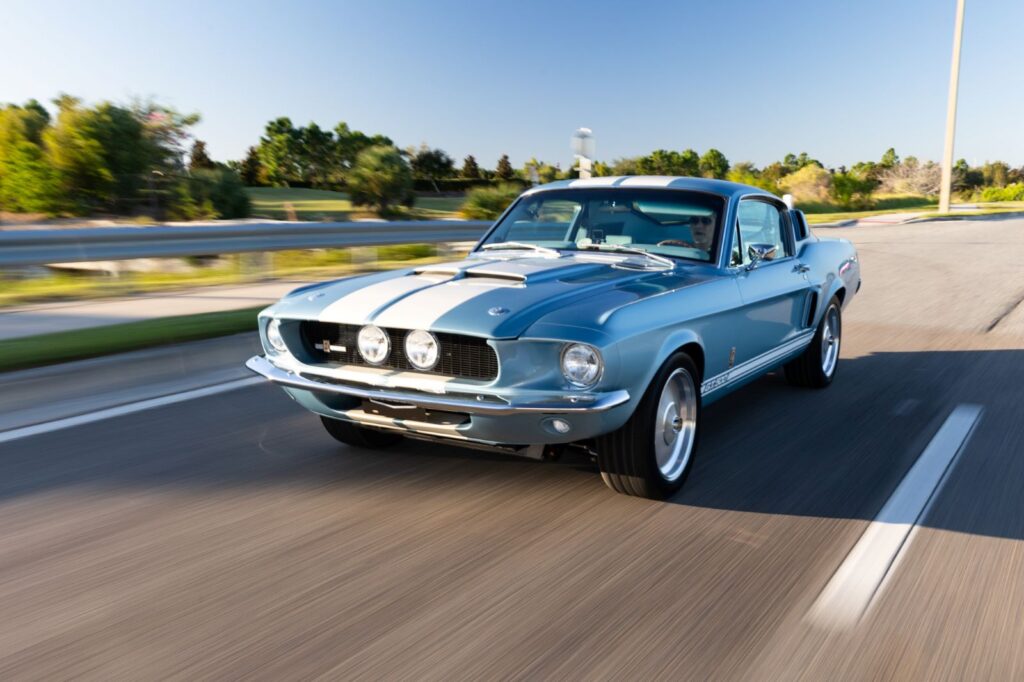 Front-quarter rolling shot of a 1967 Revology Shelby GT500 in metallic blue with white racing stripes on a scenic road.