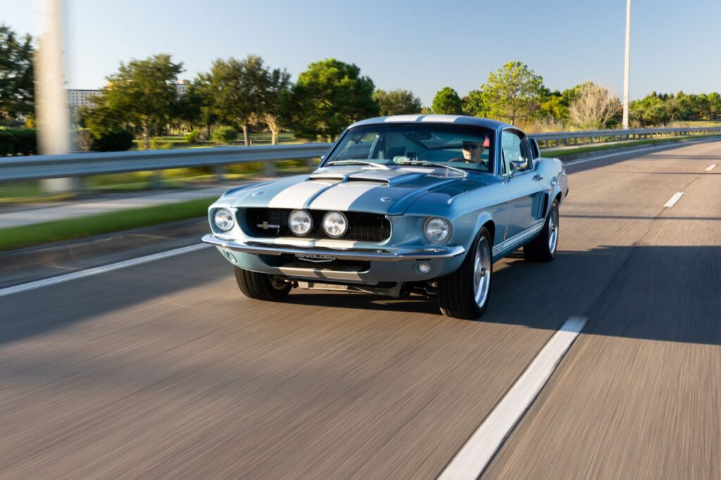 Action shot of a 1967 Revology Shelby GT500 in metallic blue with white racing stripes cruising on a multi-lane highway.