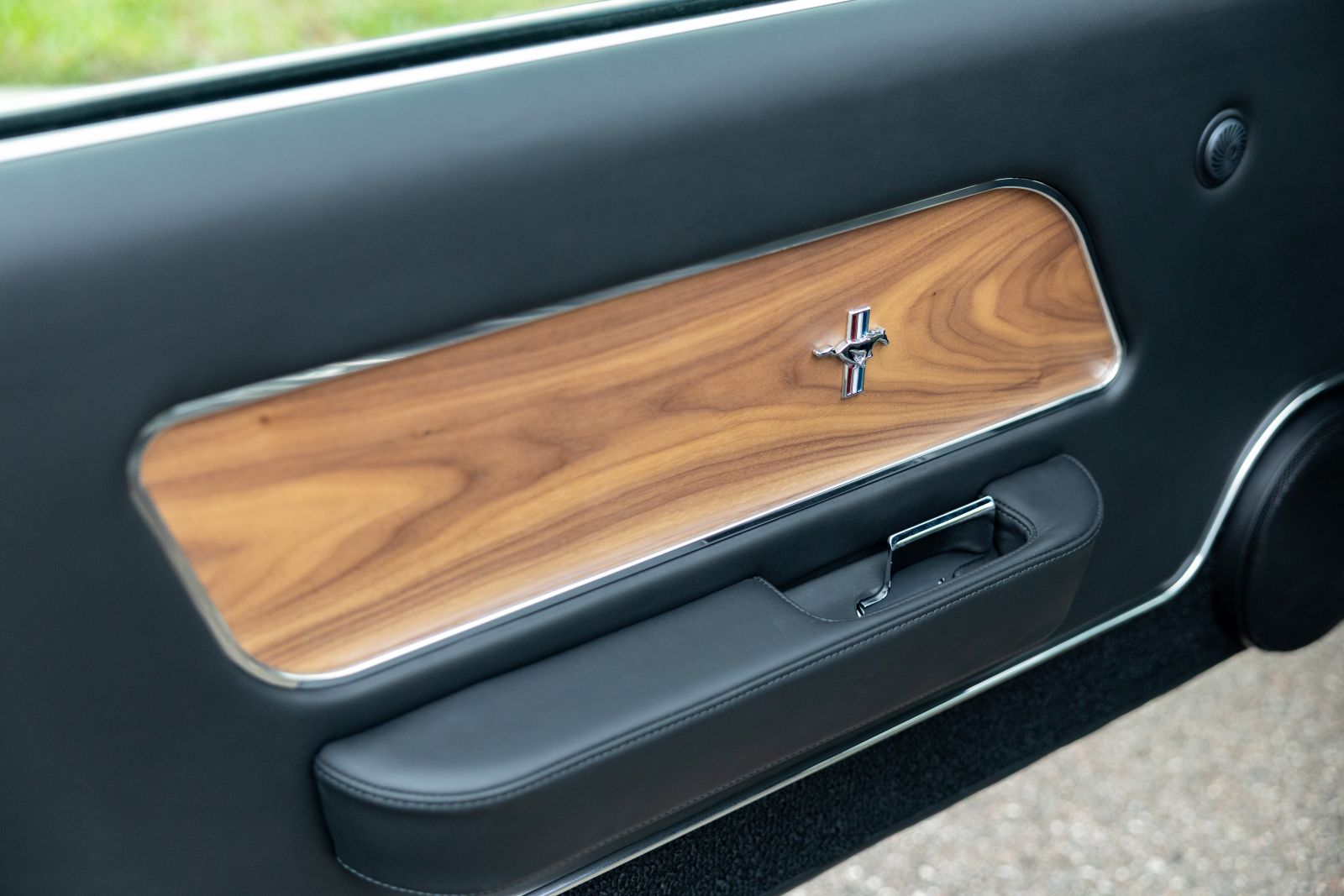 Close-up of a Revology 1969 Ford Mustang Boss 429 interior door panel featuring hand-wrapped black leather, a genuine Black American Walnut wood veneer insert, and a chrome Mustang "Running Horse" emblem.