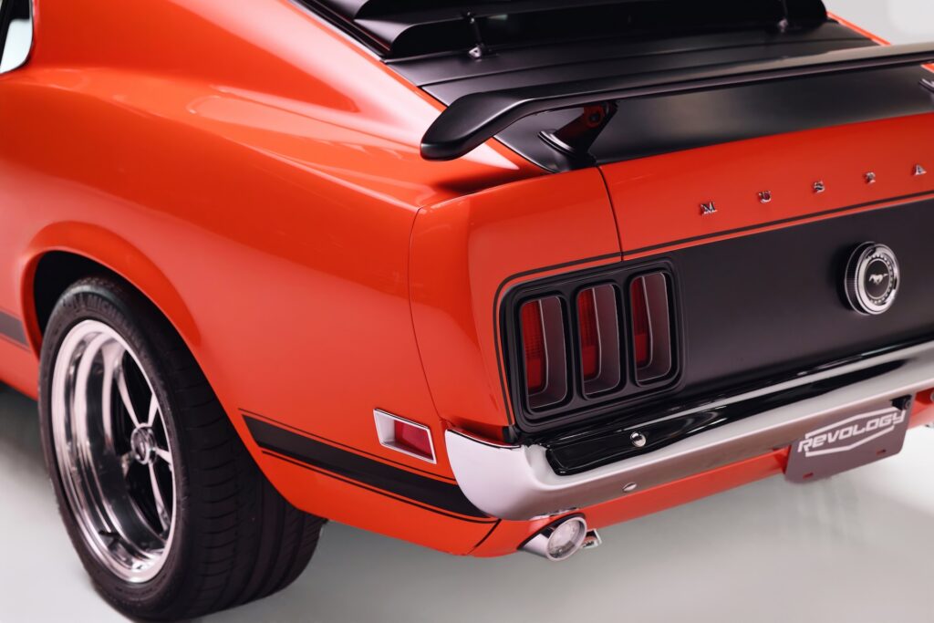 Close-up of the rear left quarter of an orange Revology 1970 Ford Mustang Boss 302, featuring the black honeycomb tail panel, LED tri-bar taillights, rear window louvers, decklid spoiler, and a 17-inch Magnum 500-style wheel.
