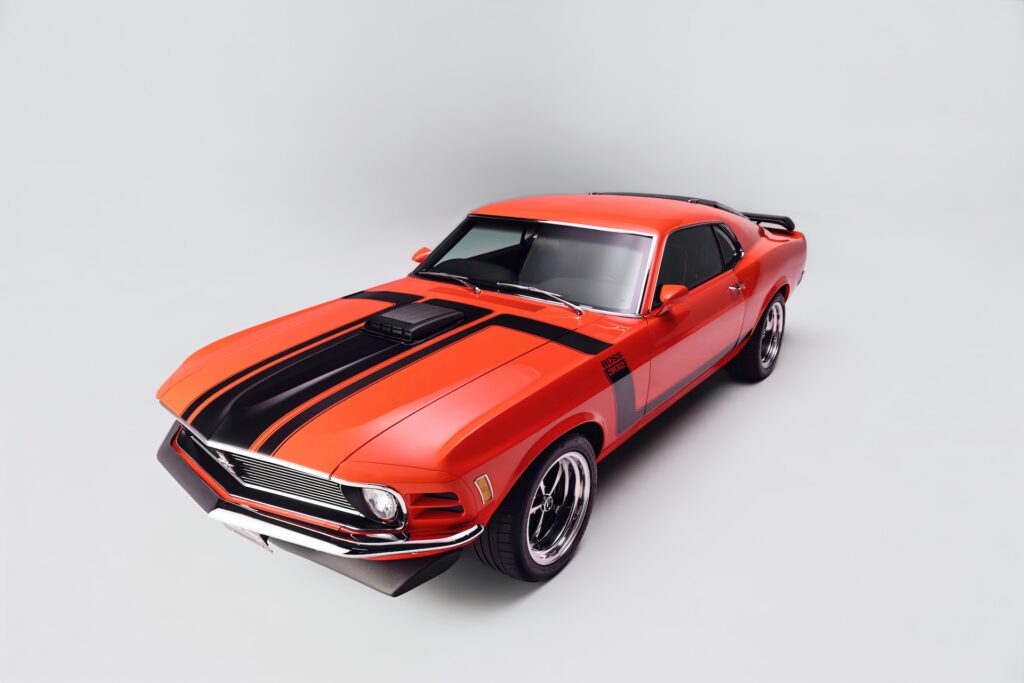 High-angle front three-quarter view of a red Revology 1970 Ford Mustang Boss 302 restomod, featuring a black shaker hood scoop, hockey stick side stripes, rear window louvers, and 17-inch Magnum 500 wheels on a white studio background.