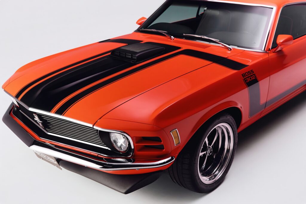 High-angle front three-quarter view of a red Revology 1970 Ford Mustang Boss 302 restomod, featuring matte black hood stripes, a functional shaker hood scoop, and 17-inch Magnum 500 wheels on a white background.