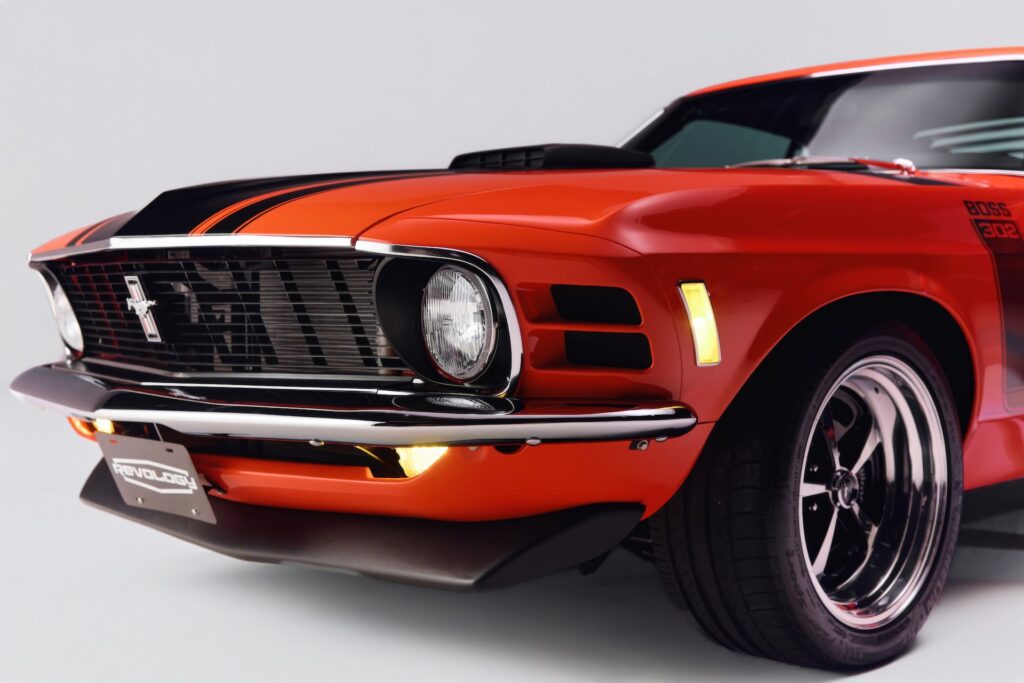 Close-up of the front-left fascia of an orange Revology 1970 Ford Mustang Boss 302 restomod, featuring illuminated LED marker lights, the iconic 1970 "shark fin" grille, a functional shaker hood scoop, and 17-inch Magnum 500-style wheels.