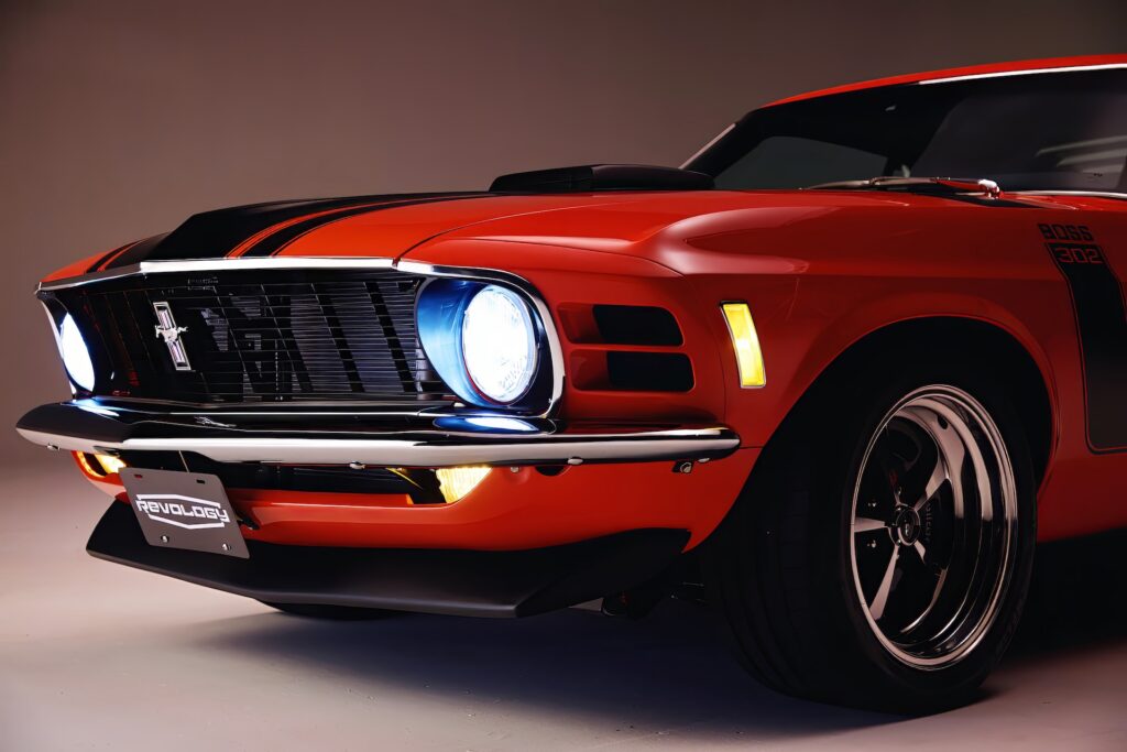 Close-up of an orange Revology 1970 Ford Mustang Boss 302 with illuminated LED headlights and amber marker lights, featuring the 1970-specific "shark fin" grille, a functional shaker hood scoop, and 17-inch Magnum 500 wheels.