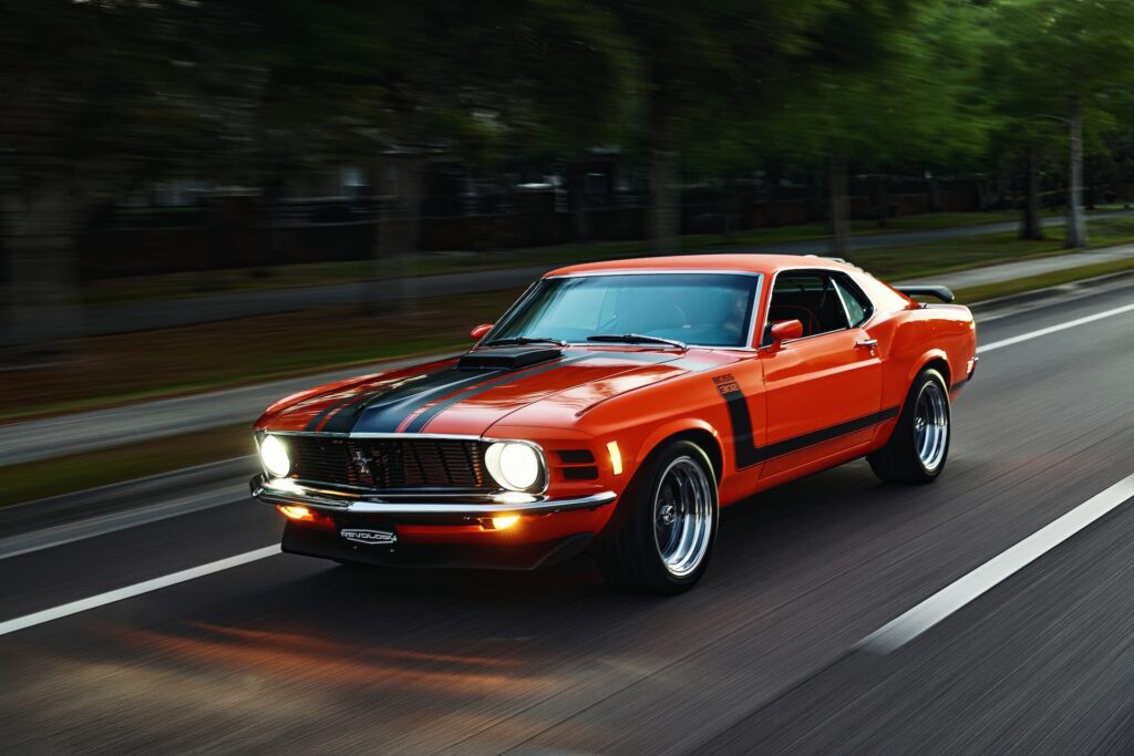 Front three-quarter motion shot of an orange Revology 1970 Ford Mustang Boss 302 restomod with LED headlights illuminated, showcasing the shaker hood scoop and black hockey stick stripes while accelerating on an asphalt road.