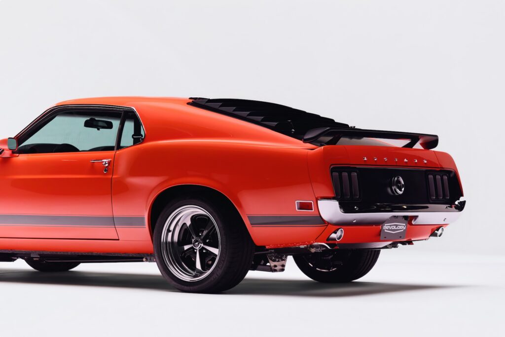 Rear three-quarter view of a vibrant red Revology 1970 Ford Mustang Boss 302 restomod, showcasing the 1970-exclusive blackout honeycomb tail panel, rear window louvers, decklid spoiler, and 17-inch Magnum 500 wheels.