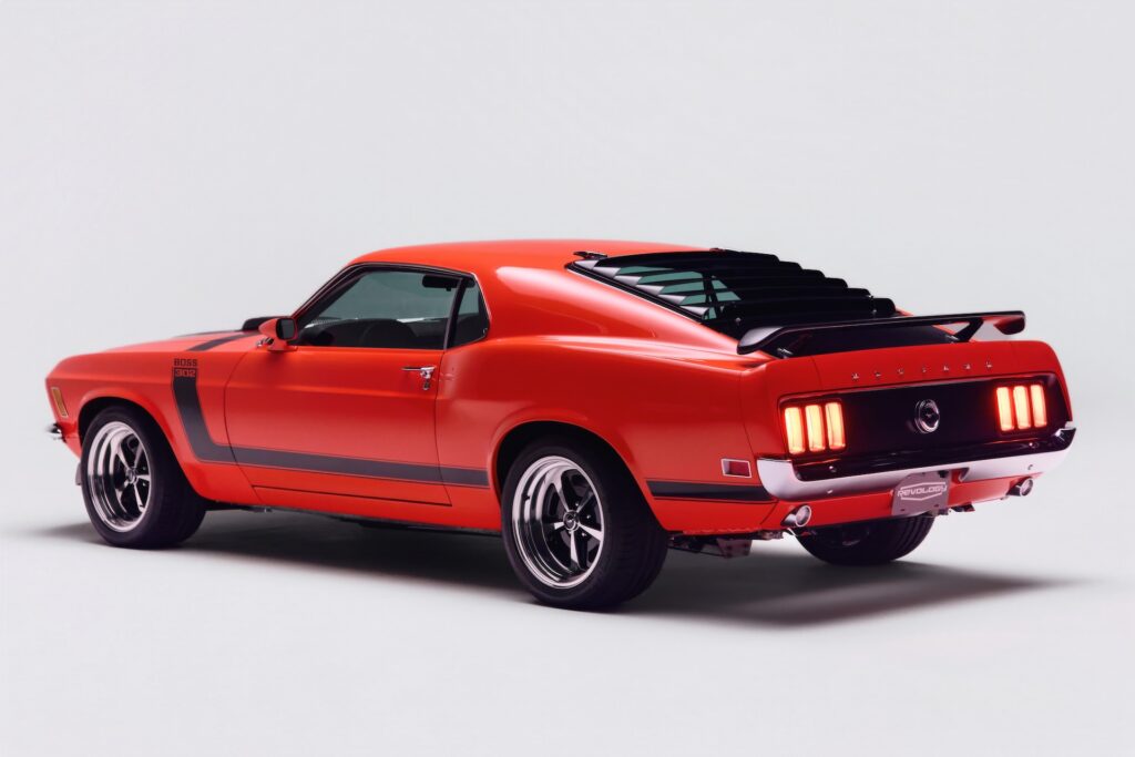 Rear three-quarter view of a red Revology 1970 Ford Mustang Boss 302 restomod with illuminated LED tri-bar taillights, black rear window louvers, decklid spoiler, and 17-inch Magnum 500 wheels.