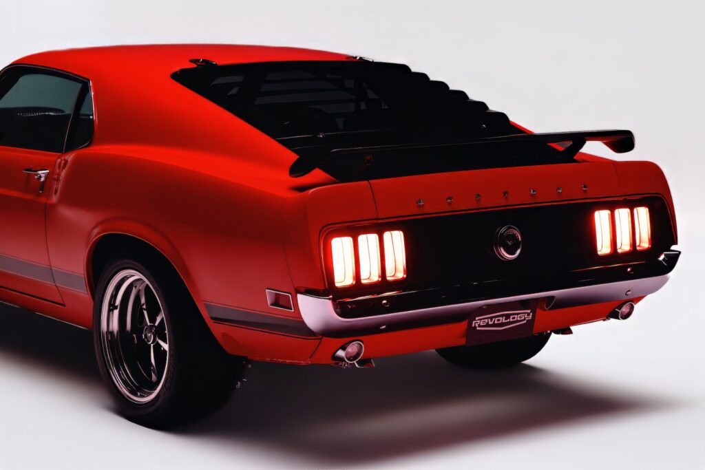 Rear three-quarter view of a red Revology 1969 Ford Mustang fastback restomod featuring illuminated sequential LED tri-bar taillights, black rear window louvers, and a pedestal spoiler.