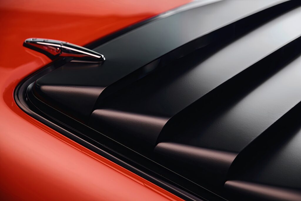 Close-up detail of the matte black rear window louvers (Sport Slats) on a red Revology 1970 Boss 302 Mustang, highlighting the chrome-plated mounting hardware and the contrast between the textured black slats and vibrant orange-red body paint.