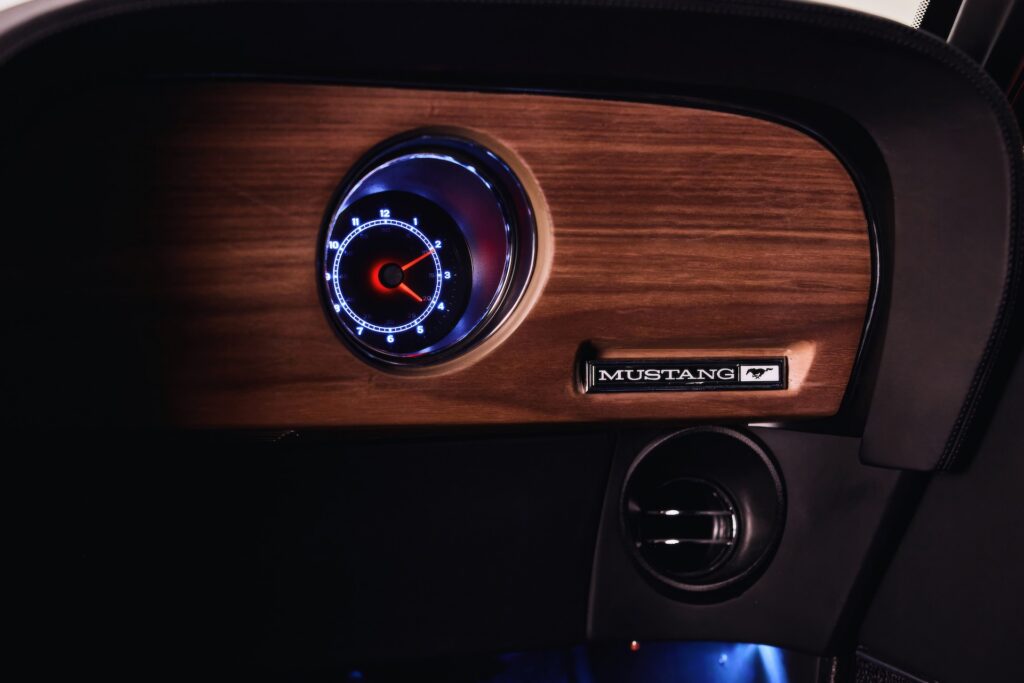 Close-up photograph of the passenger-side dashboard in a customized classic Ford Mustang restomod. The view showcases a dark wood grain veneer panel, a singular circular analog clock gauge with modern blue and white LED backlighting and a red second hand, a "MUSTANG" text script with running horse emblem, a modern directional A/C vent, and ambient blue footwell lighting below.