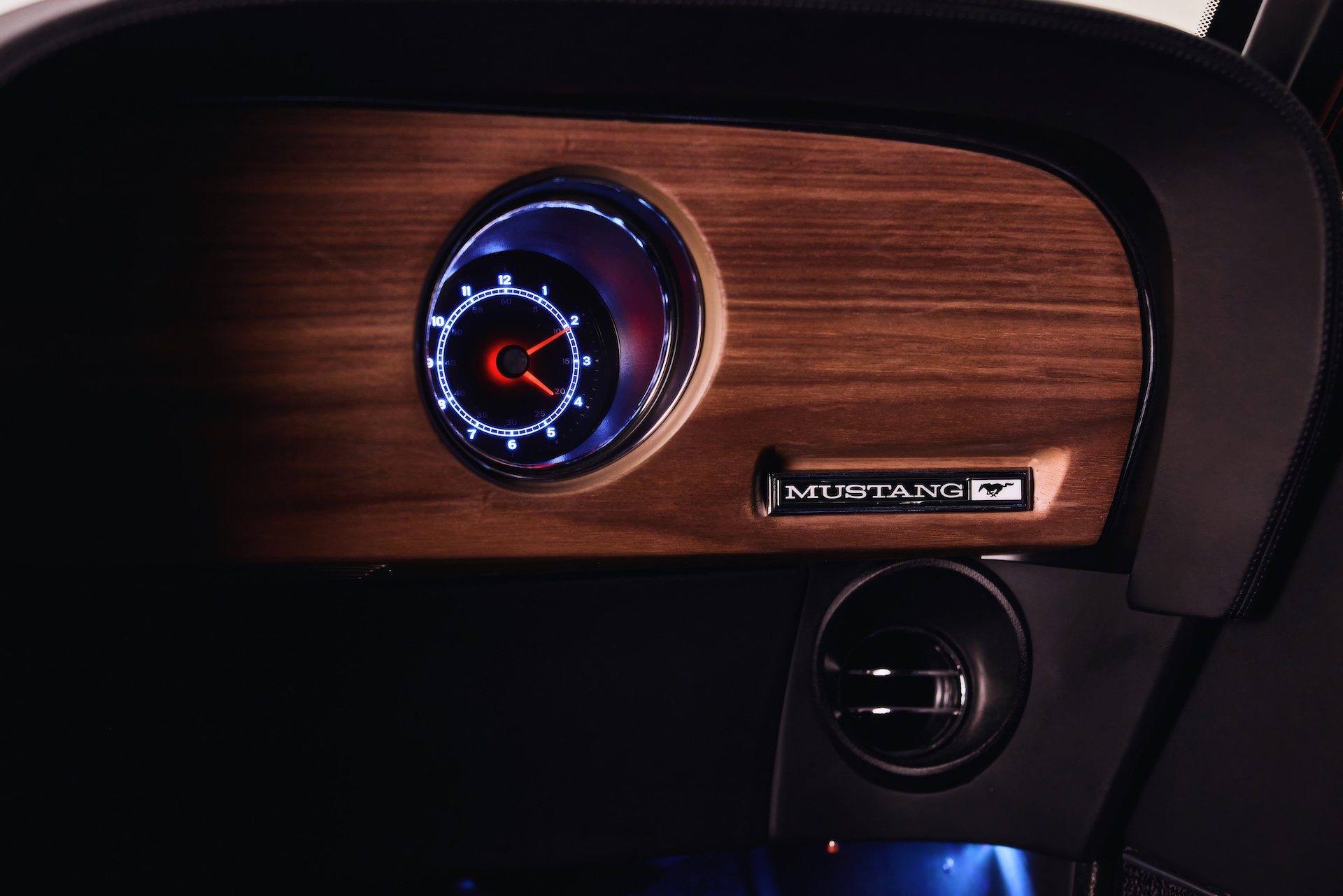 Close-up photograph of the passenger-side dashboard in a customized classic Ford Mustang restomod. The view showcases a dark wood grain veneer panel, a singular circular analog clock gauge with modern blue and white LED backlighting and a red second hand, a "MUSTANG" text script with running horse emblem, a modern directional A/C vent, and ambient blue footwell lighting below.