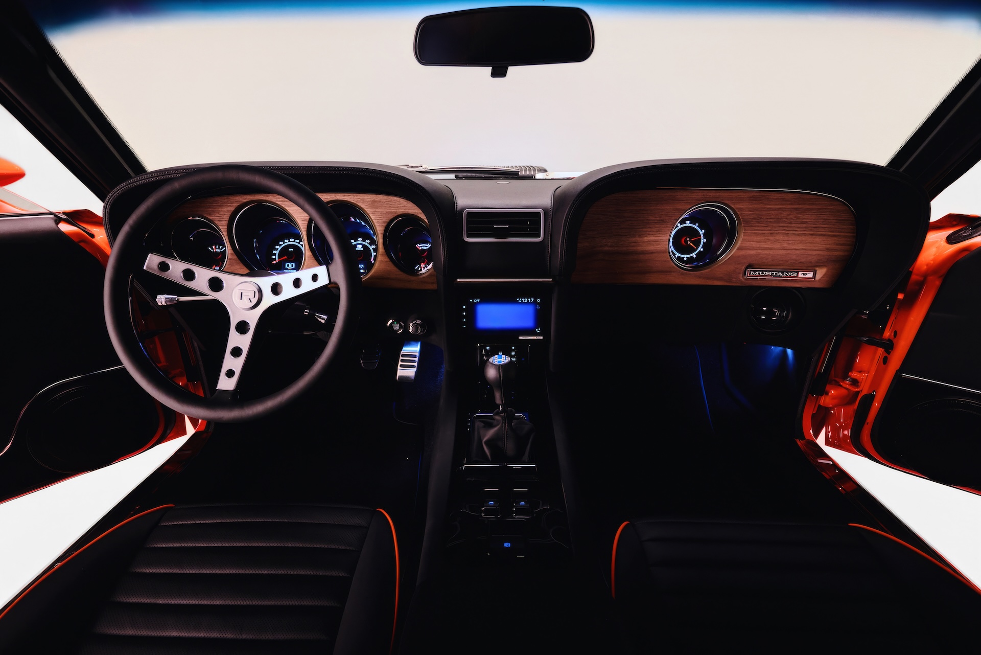 Wide-angle interior of a Revology 1969 Mustang restomod featuring an active 7-inch touchscreen infotainment system, walnut wood grain dashboard, and black leather performance seats with orange contrast piping.