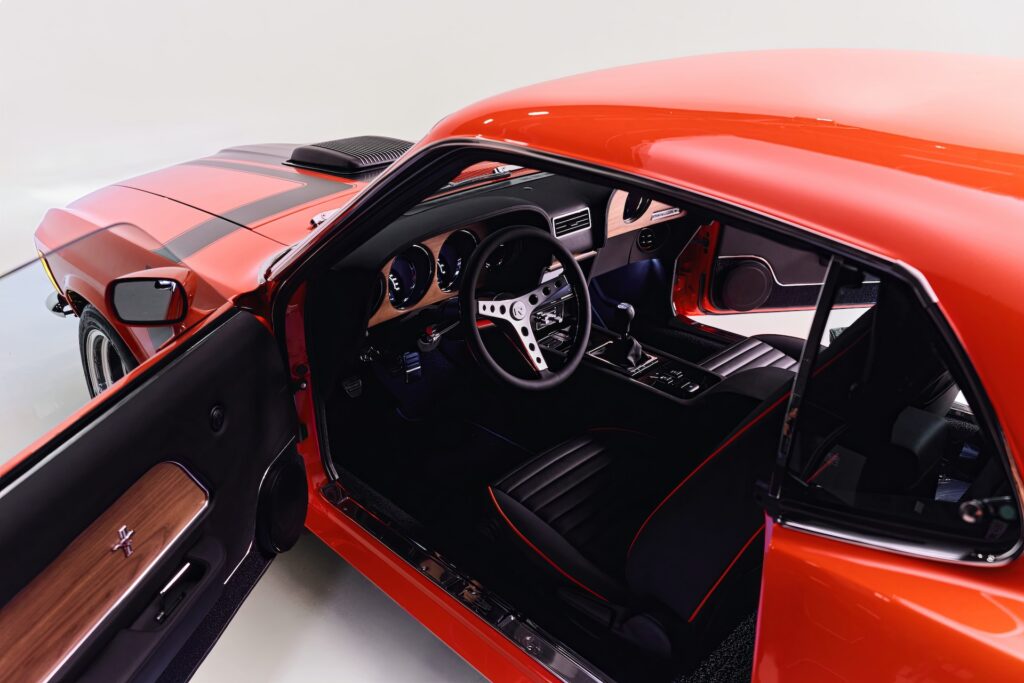 Interior view of a Revology 1970 Ford Mustang Boss 302 restomod, featuring black leather performance seats with red stitching, a walnut wood veneer dashboard, a 3-spoke aluminum steering wheel, and a modern 6-speed manual shifter.