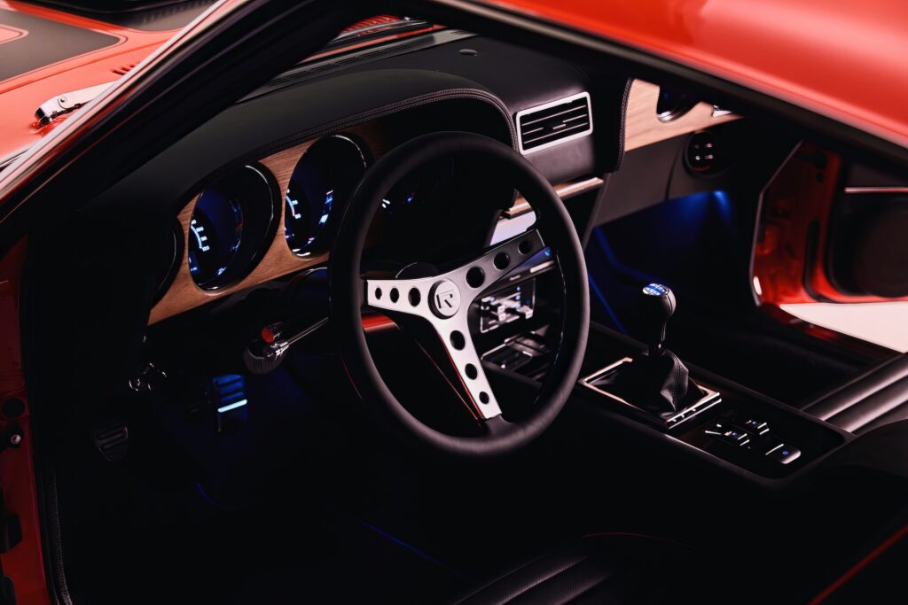 Interior cockpit view of a red Revology 1970 Ford Mustang Boss 302 restomod, featuring blue LED-backlit gauges, a three-spoke aluminum steering wheel, genuine walnut wood dashboard trim, and a 6-speed manual shifter.