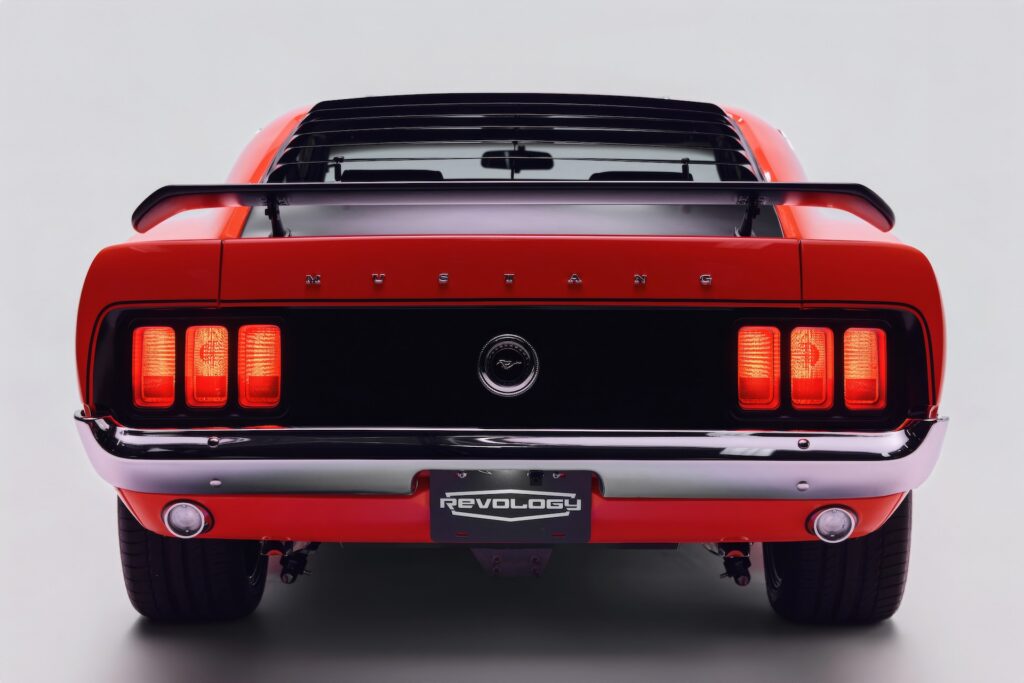 Straight-on rear view of a vibrant red Revology 1970 Ford Mustang Boss 302 restomod with illuminated LED tri-bar taillights, black honeycomb tail panel, rear window louvers, and a pedestal-mounted spoiler.