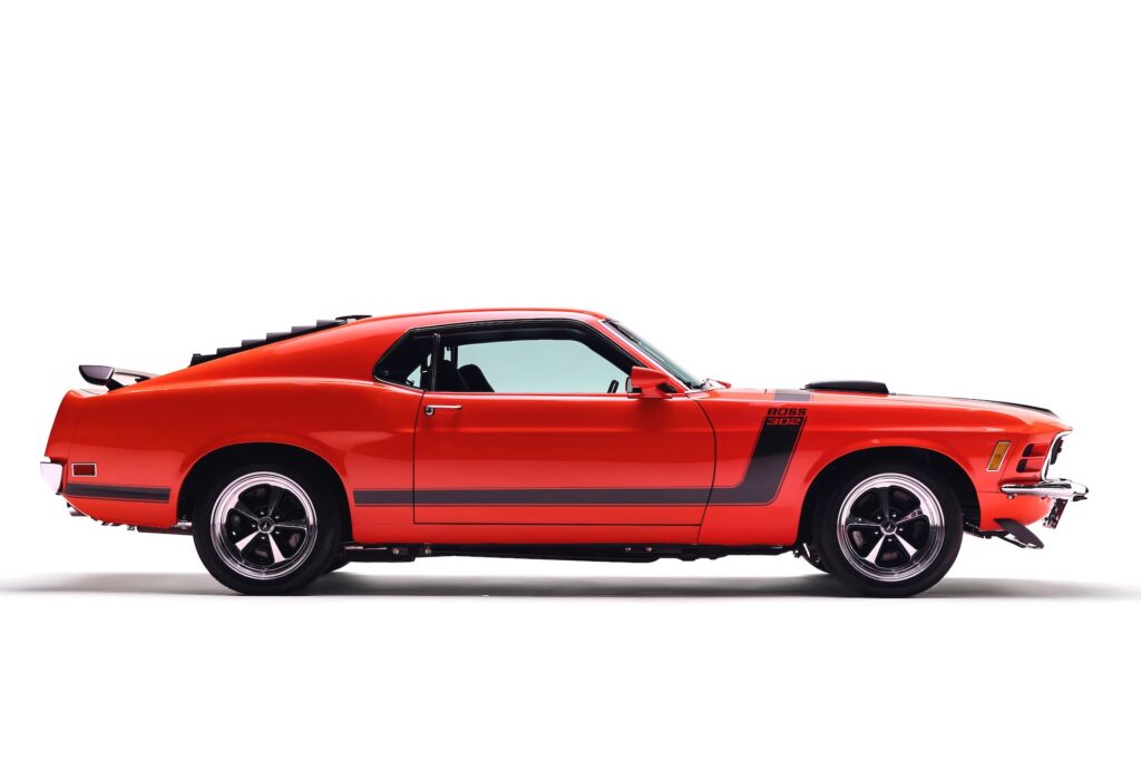 Full side profile of a Revology 1970 Ford Mustang Boss 302 restomod in vibrant red, featuring black hockey stick side stripes, rear window louvers, a rear decklid spoiler, and modern 17-inch Magnum 500 wheels.
