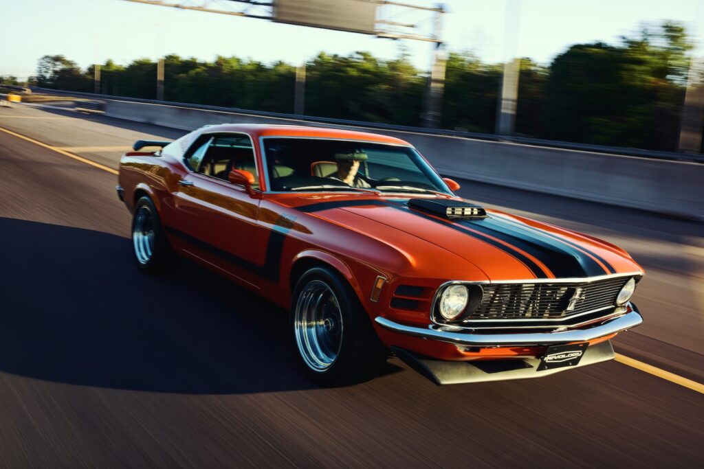 High-angle front three-quarter motion shot of an orange Revology 1970 Ford Mustang Boss 302 restomod on a highway, highlighting the functional shaker hood scoop, front chin spoiler, and larger 17-inch Magnum 500-style wheels.
