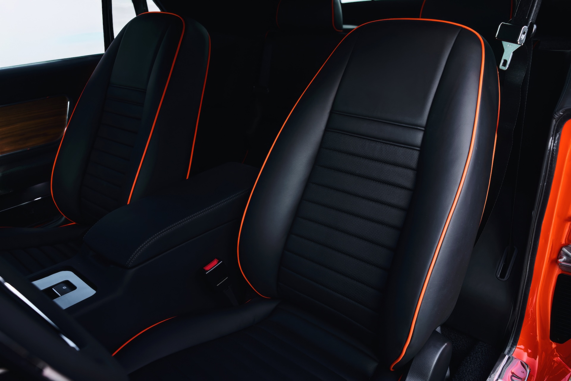 Close-up interior photograph of custom high-back bucket seats in a classic Ford Mustang restomod (late 1960s style). The seats are upholstered in premium black leather with bright orange contrast piping and vertical perforated inlays.