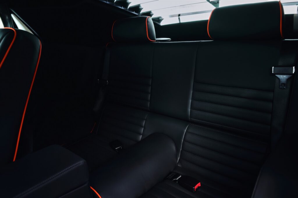 Close-up of the rear passenger seats in a Revology 1970 Ford Mustang Boss 302, featuring hand-wrapped black leather, horizontal pleats, and custom red contrast piping on the headrests and bolsters.