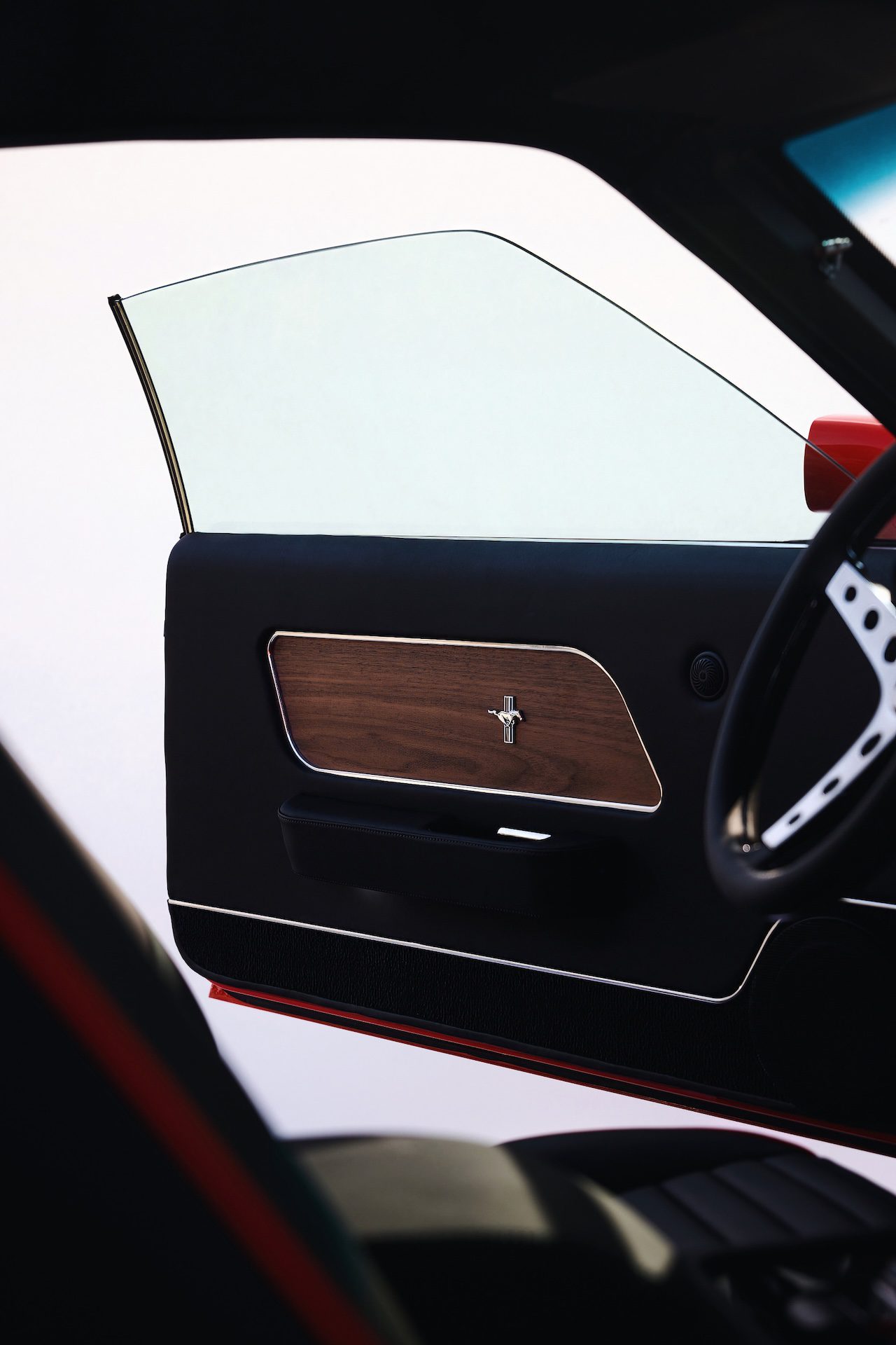 Interior door panel of a Revology classic Mustang restomod featuring premium black leather, a walnut-style wood grain insert, and a chrome Mustang "Running Horse" emblem.