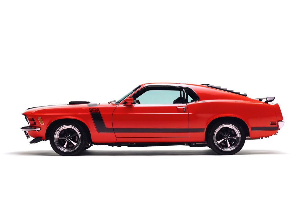 Full side profile of a red Revology 1970 Ford Mustang Boss 302 restomod featuring black hockey stick side stripes, rear window louvers, a rear spoiler, and 17-inch Magnum 500 wheels.
