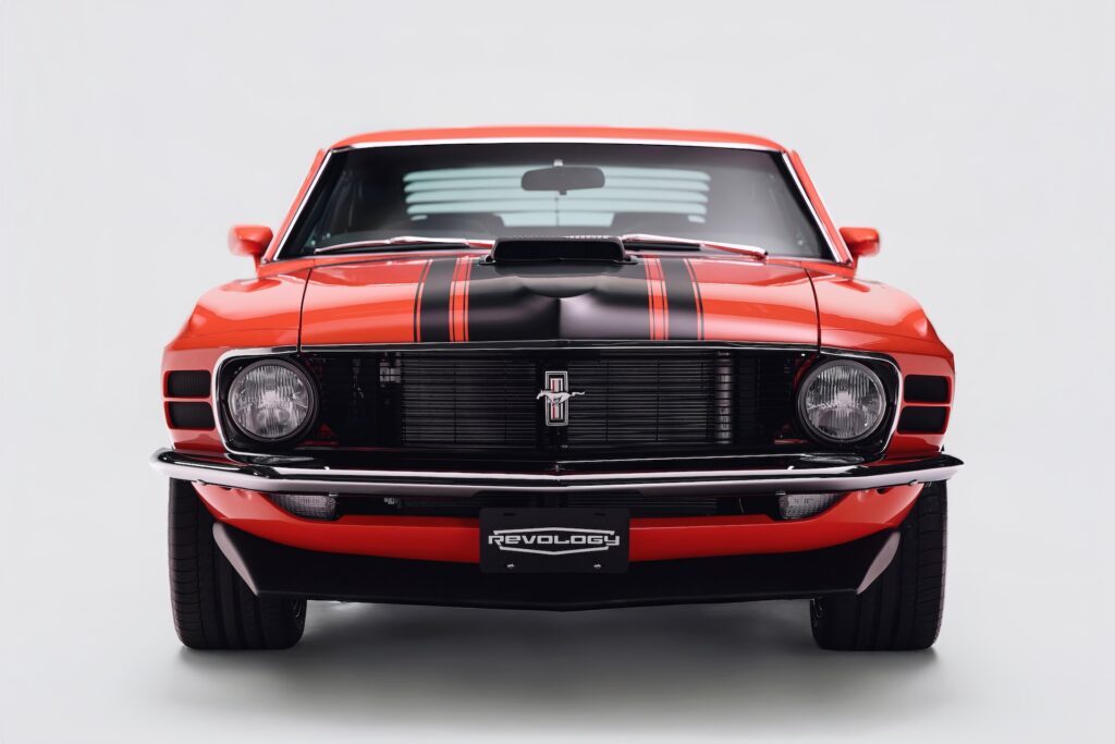 Head-on studio portrait of a red Revology 1970 Ford Mustang Boss 302 restomod, showcasing the 1970-specific dual-headlight grille, black hood stripes, functional shaker scoop, and aggressive black chin spoiler.