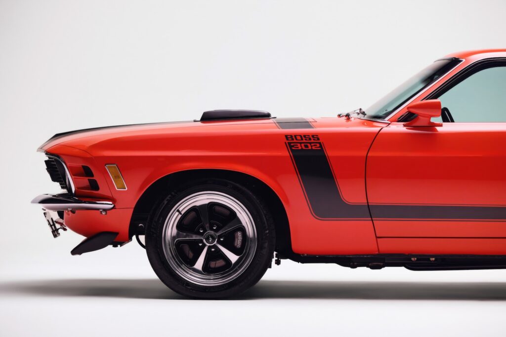 Side profile close-up of the front fender on an orange Revology 1970 Ford Mustang Boss 302, featuring black hockey stick stripes, BOSS 302 nomenclature, a shaker hood scoop, and 17-inch Magnum 500 wheels with visible performance brakes.