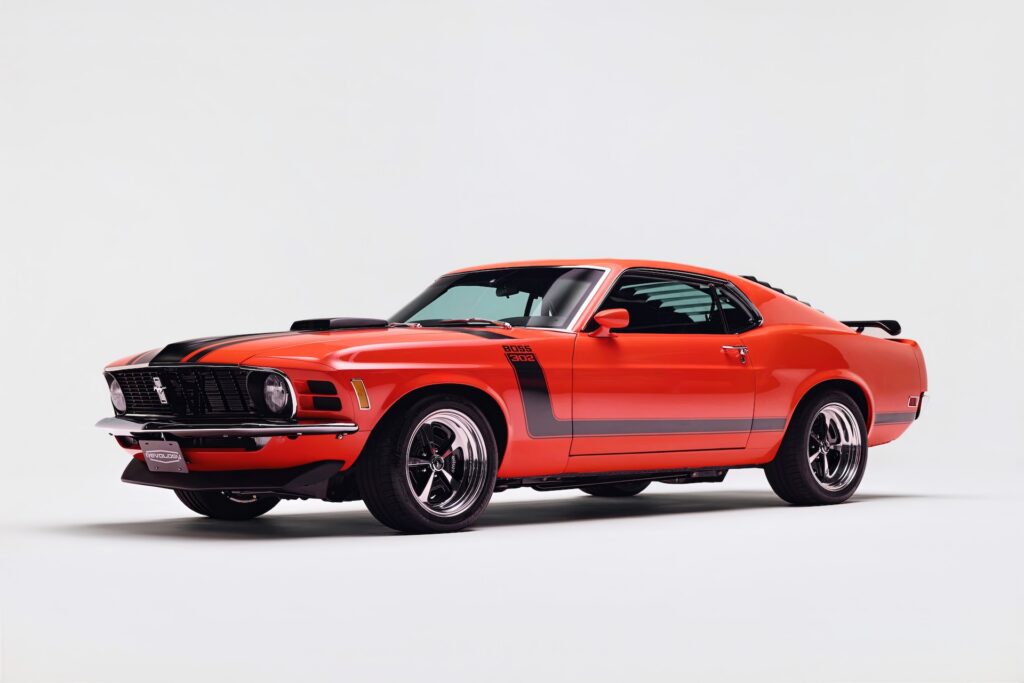 Front three-quarter view of a Revology 1970 Ford Mustang Boss 302 in vibrant red, featuring black hockey stick stripes, a functional shaker hood, 17-inch Magnum 500 wheels, and rear window louvers.